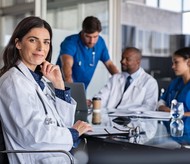 Portrait of mature female doctor sitting in meeting room with specialist and nurses discussing case in background. Successful woman doctor in labcoat and stethoscope in hospital. Smiling pediatrician looking at camera with medical staff brainstorming